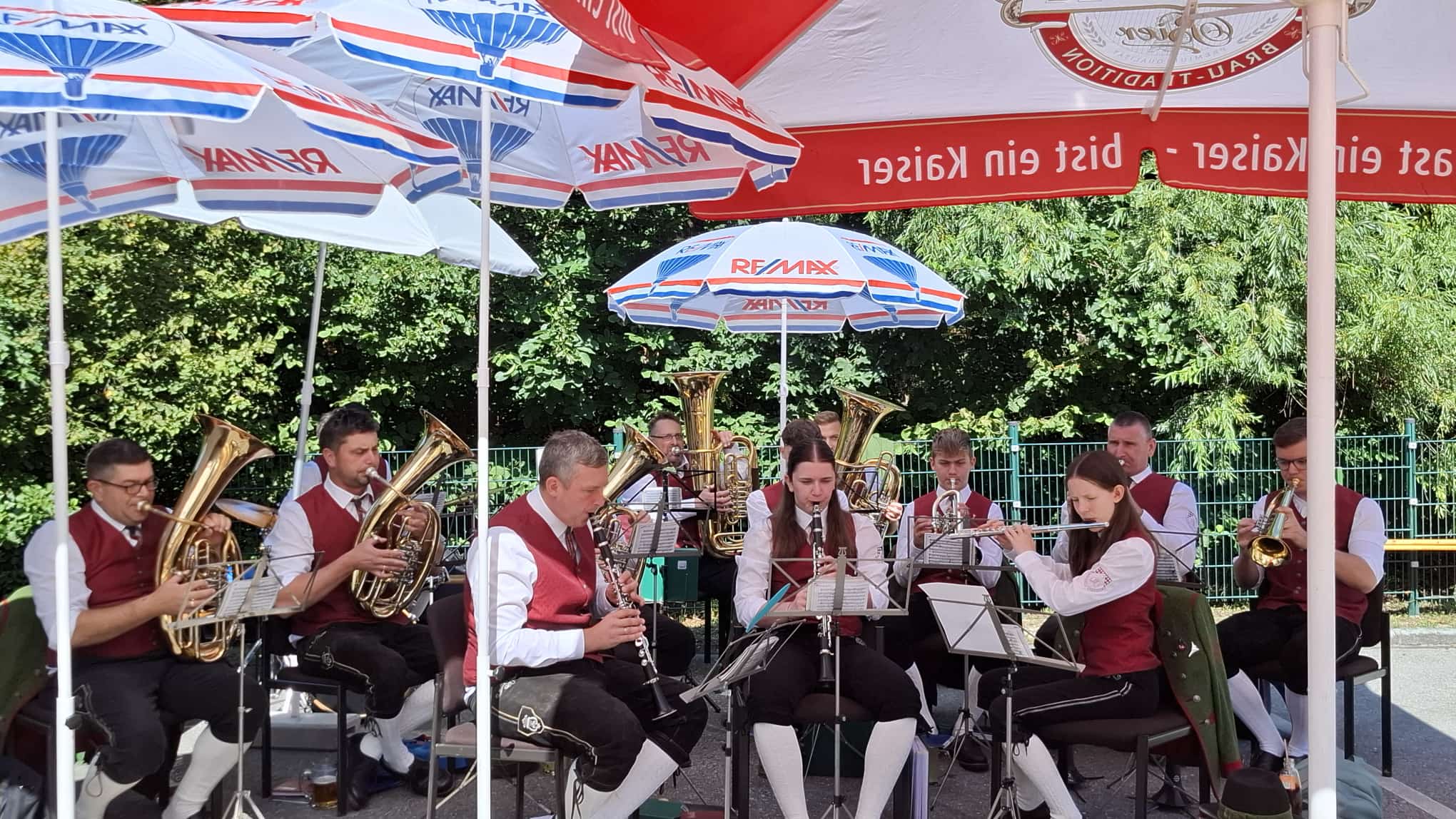 Blasmusikgruppe spielt unter roten und blauen Sonnenschirmen, Musiker in Tracht
