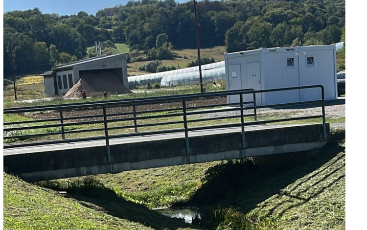 Brücke über Bach mit Gewächshäusern und Container-Gebäude im Hintergrund