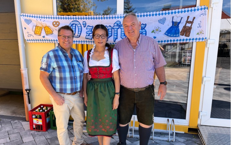 O'zapft is: Mann in Tracht und zwei weitere Personen vor Bierkisten und Fahne