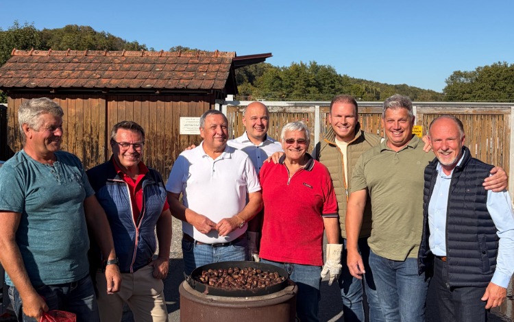 Acht Männer stehen vor einem Holzhaus und einem Zaun, im Vordergrund ein Rost mit Maroni.