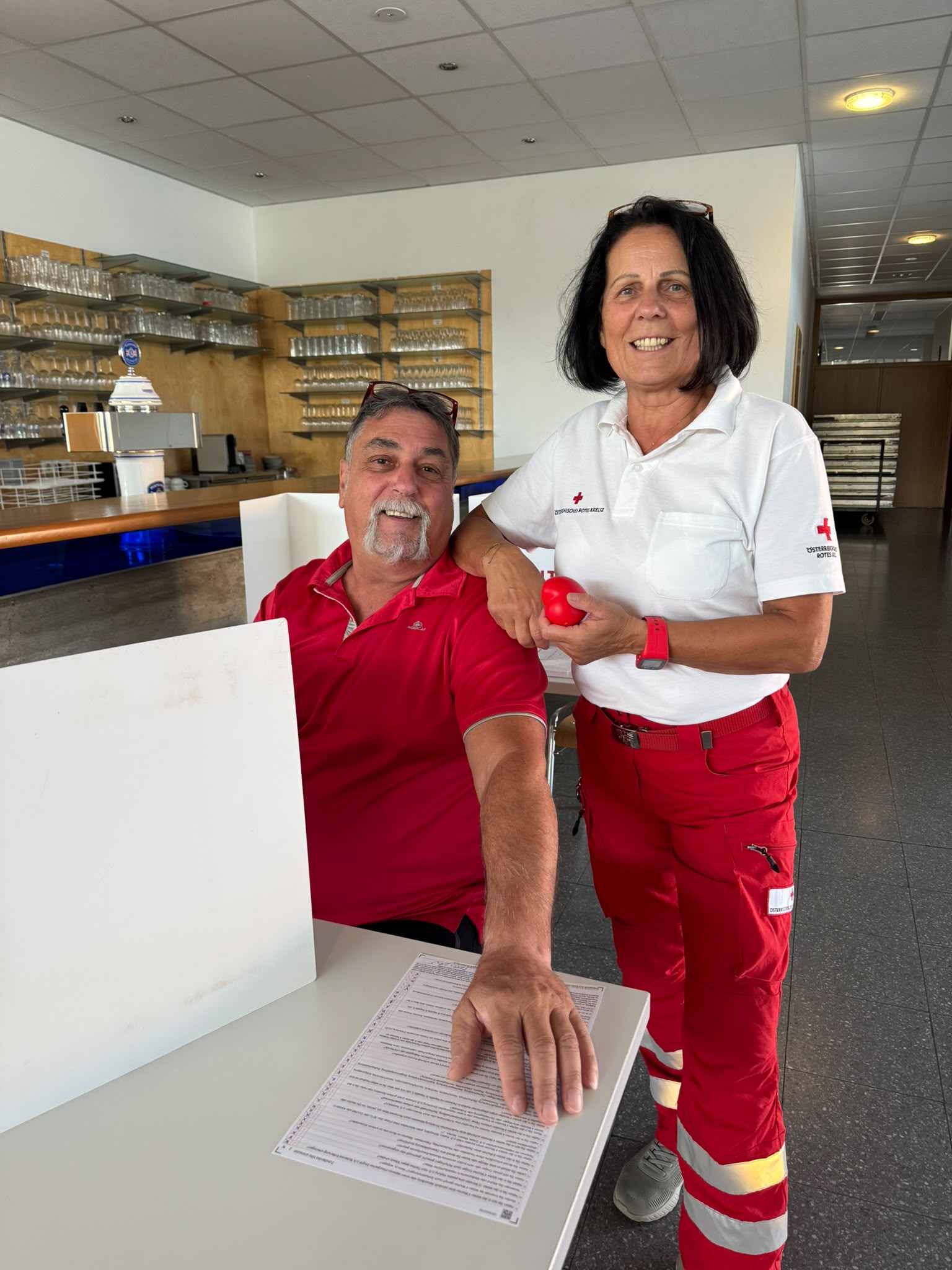 Rotes Kreuz Mitarbeiterin mit rotem Ball und Mann in rotem Poloshirt an einem Tisch mit Formular